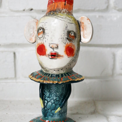 Ceramic figure with a stylized face and colorful attire against a white brick wall.