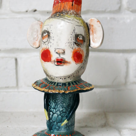 Ceramic figure with a stylized face and colorful attire against a white brick wall.